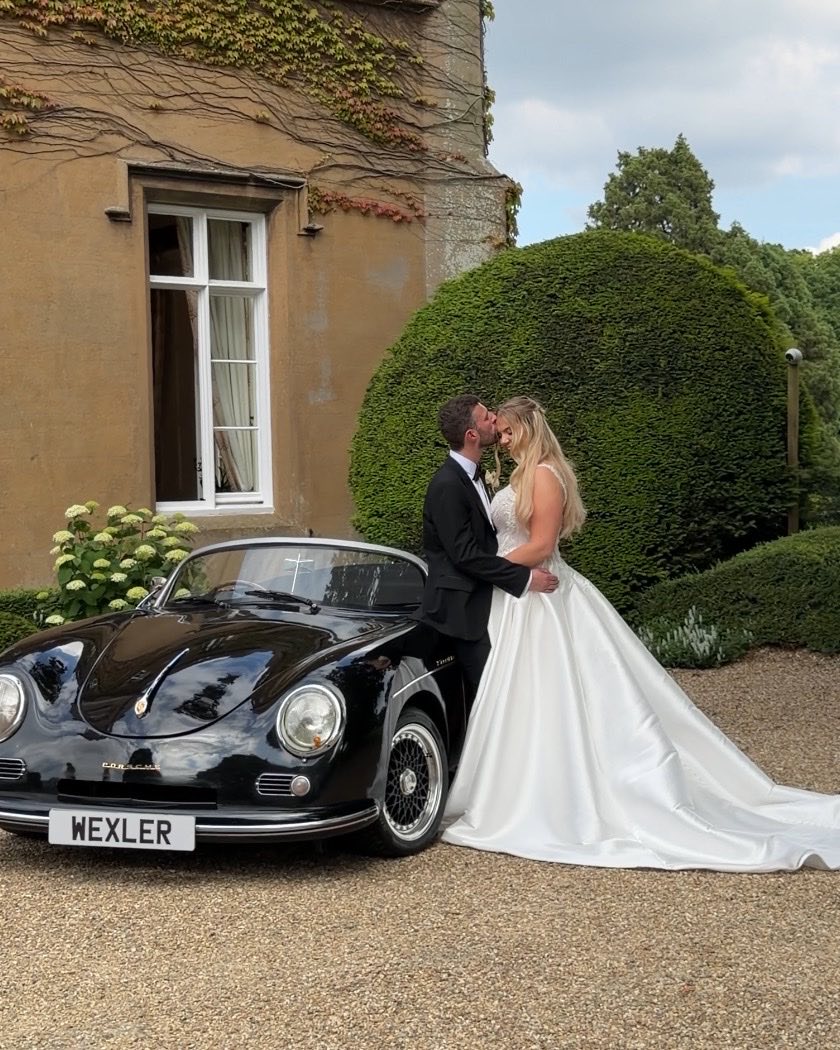 Newlyweds kissing beside a classic car outside a country house venue